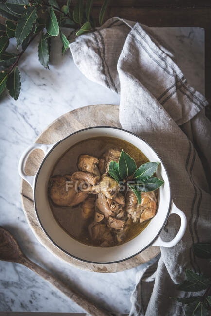 Top view of stewed chicken with broth in pan decorated with green leaves on white marble table — Stock Photo