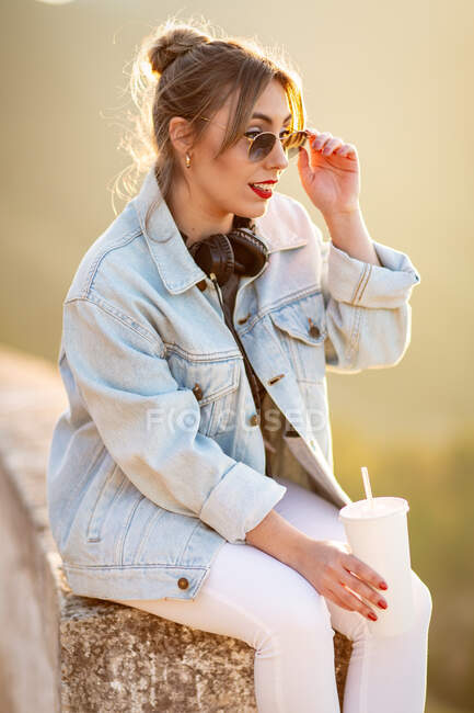 Vista lateral da jovem mulher em desgaste casual com óculos de sol descansando em cerca rochosa com bebida e ouvir música com fones de ouvido — Fotografia de Stock