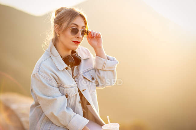 Vue latérale de la jeune femme en tenue décontractée avec des lunettes de soleil reposant sur une clôture rocheuse avec des boissons et écoutant de la musique avec écouteurs — Photo de stock