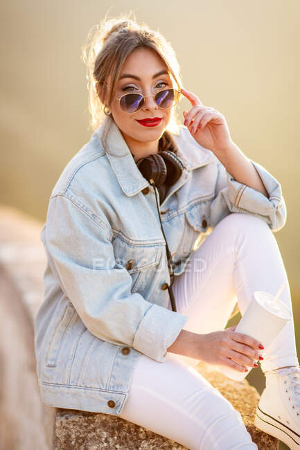 Задоволена блондинка в модних сонцезахисних окулярах і повсякденний одяг, сидячи на кам'янистому паркані і дивлячись на камеру на сонячному світлі на розмитому фоні — стокове фото