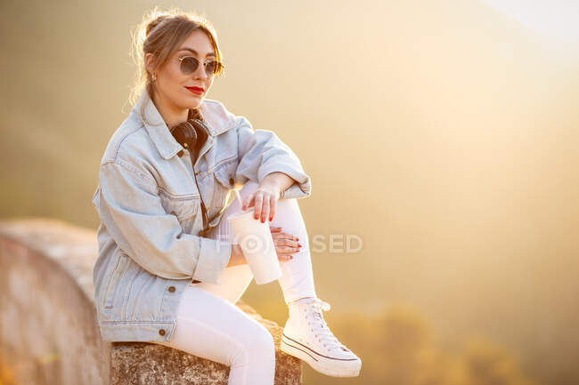 Satisfied blond haired lady in trendy sunglasses and casual wear sitting on rocky fence and looking at camera in sunlight on blurred background — Stock Photo