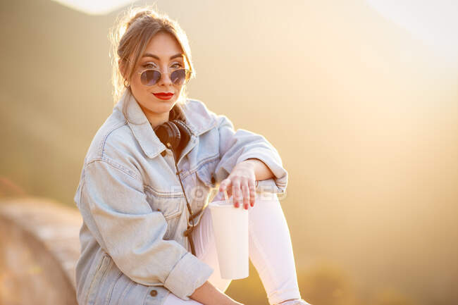 Вид збоку молодої жінки в повсякденному одязі з сонцезахисними окулярами, що спираються на кам'яний паркан з чашкою напоїв і навушниками навколо шиї — стокове фото