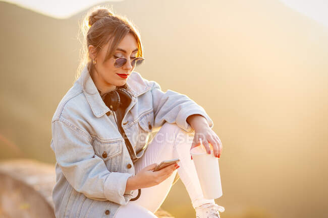 Nette Dame mit trendiger Sonnenbrille lächelt, während sie ihr Handy im warmen Sonnenlicht auf verschwommenem Hintergrund benutzt — Stockfoto