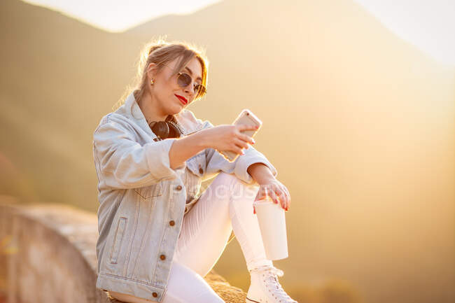 Nette Dame mit trendiger Sonnenbrille lächelt, während sie ein Selfie auf dem Handy im warmen Sonnenlicht vor verschwommenem Hintergrund macht — Stockfoto