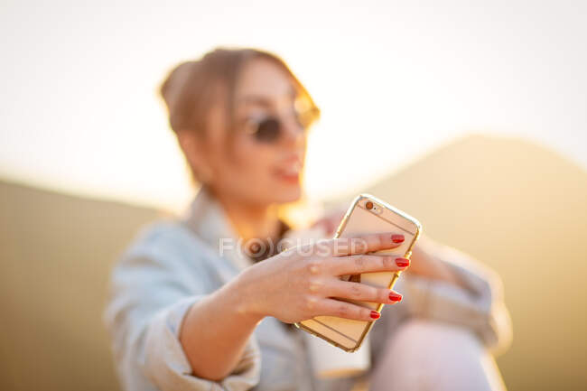 Мила леді в модних сонцезахисних окулярах посміхається, беручи селфі на мобільний телефон у тепле світло сонця на розмитому фоні — стокове фото