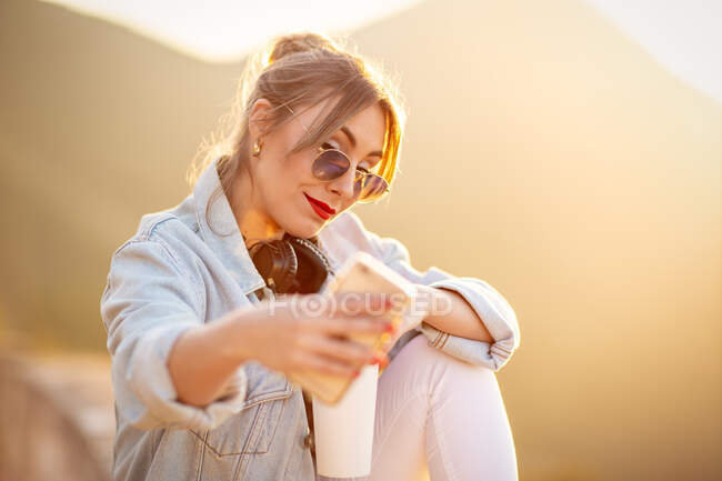 Bonita senhora na moda óculos de sol sorrindo ao tomar selfie no celular em luz quente do sol no fundo borrado — Fotografia de Stock