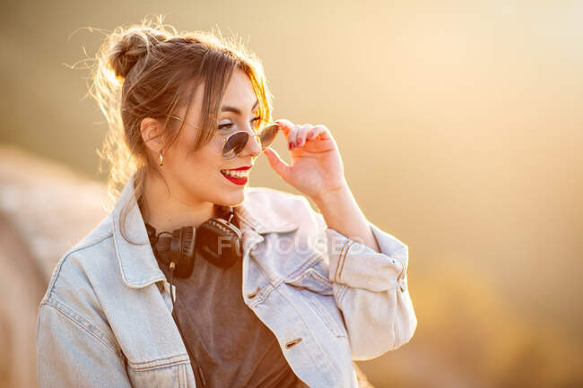 Giovane donna allegra con occhiali da sole in abito casual alla moda sorridente e guardando altrove nella giornata di sole — Foto stock
