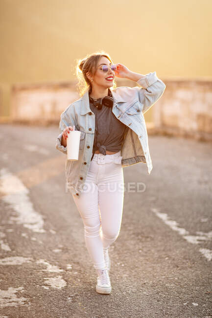 Mujer rubia alegre con un atuendo elegante y gafas de sol caminando con bebida y sonriendo sobre un fondo borroso - foto de stock