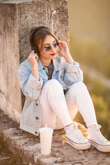 Junge Frau in Freizeitkleidung mit Sonnenbrille lehnt mit Getränken auf felsigem Zaun und hört Musik mit Kopfhörern — Stockfoto