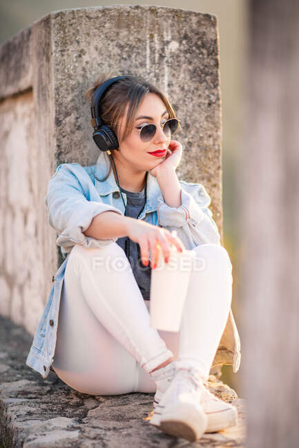 Mujer joven en ropa casual con gafas de sol descansando sobre una valla rocosa con bebida y escuchando música con auriculares - foto de stock
