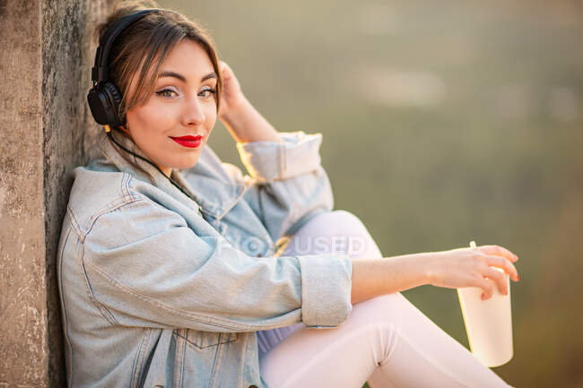 Вид сбоку на вдумчивую даму в повседневной одежде, отдыхающую на скалистом заборе с напитком и слушающую музыку с наушниками — стоковое фото