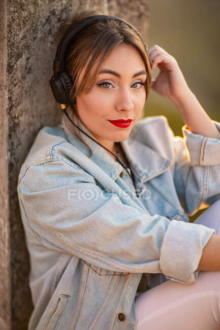 Vue latérale de la jeune femme regardant la caméra en tenue décontractée reposant sur une clôture rocheuse écoutant de la musique avec des écouteurs — Photo de stock