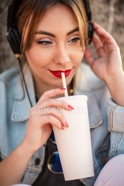 Молодая женщина в повседневной одежде отдыхает на скалистом заборе с напитком и слушает музыку в наушниках — стоковое фото