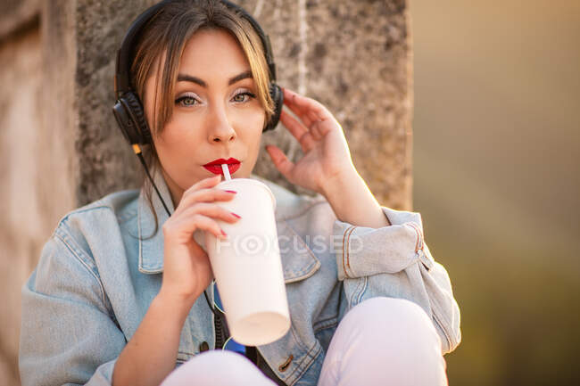 Молода жінка в повсякденному одязі відпочиває на кам'янистому паркані з напоєм і слухає музику з навушниками — стокове фото