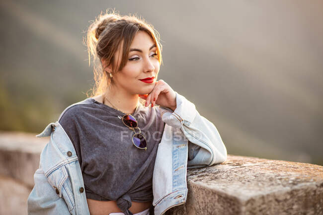 Радостная молодая женщина в модной повседневной одежде улыбается и смотрит в сторону в солнечный день — стоковое фото