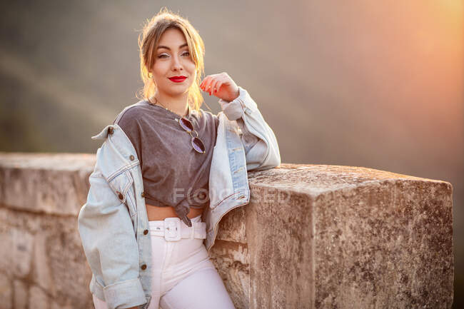 Радостная молодая женщина в модной повседневной одежде улыбается и смотрит в камеру в солнечный день — стоковое фото