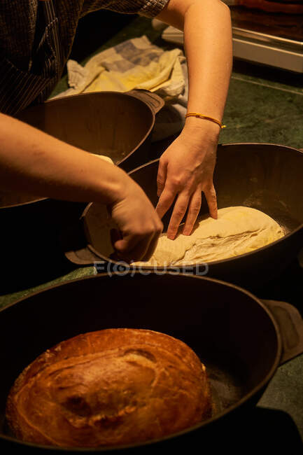 Cropped unrecognizable person hands preparing bread near cooked golden bread loaf in cast iron form on table — Stock Photo