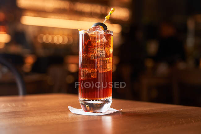 Highball glass with red alcohol cocktail with ice cubes decorated with stick with black olive on wooden counter — Stock Photo