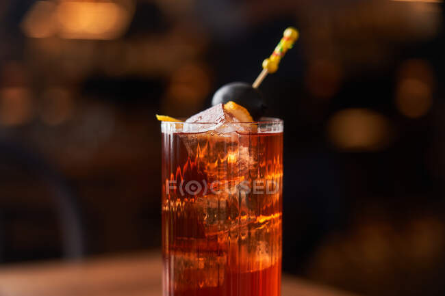 Highball glass with red alcohol cocktail with ice cubes decorated with stick with black olive on wooden counter — Stock Photo