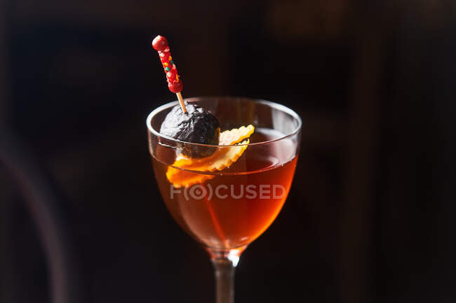 Cóctel de alcohol rojo Manhattan adornado con cereza y ralladura naranja en palo sobre fondo oscuro - foto de stock