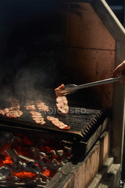 Crop chef girando correas de tocino jugoso a la parrilla mientras cocina con humo en el estante en el jardín - foto de stock