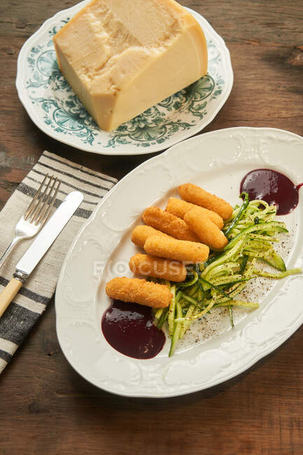 Vista superior de palitos de queso frito con pepino en rodajas y salsa barbacoa en plato sobre mesa de madera - foto de stock