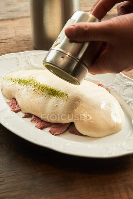 From above of faceless chef adding condiment on thick sauce covering slices of roast beef on plate — Stock Photo