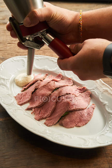 Зверху безликий шеф-кухар додає пухнастий соус на тарілці зі скибочками ніжного смаженого м'яса яловичини на дерев'яному столі — стокове фото