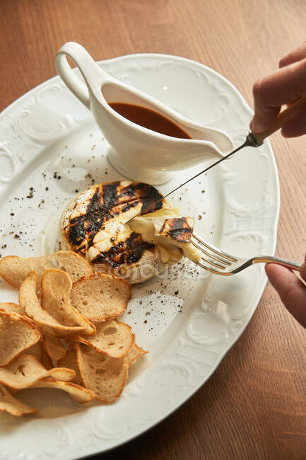From above unrecognizable person holding cutlery to eat delicious grilled chicken burger with crispy baked bread chips and BBQ sauce on plate on table — Stock Photo
