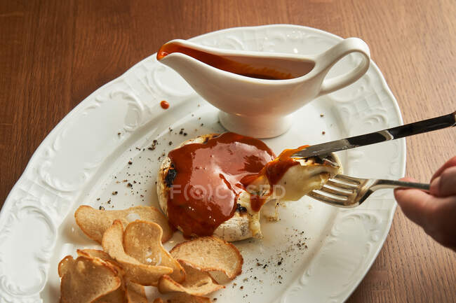 De cima pessoa irreconhecível segurando talheres para comer delicioso hambúrguer de frango grelhado com batatas fritas crocantes pão cozido e molho de churrasco no prato na mesa — Fotografia de Stock