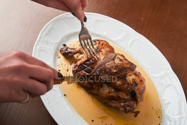 Сверху анонимная женщина держит столовые приборы и режет курицу на тарелке на столе — стоковое фото