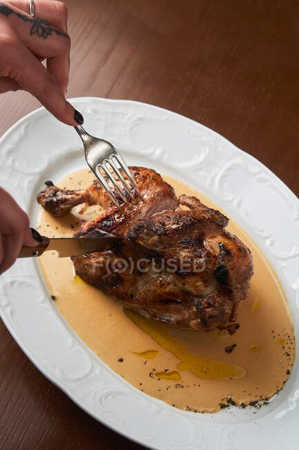 Von oben hält anonyme Frau Besteck in der Hand und schneidet gegrilltes Hühnchen auf dem Teller auf den Tisch — Stockfoto