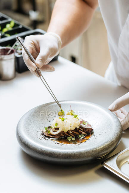 Desde arriba cocinero profesional irreconocible en guantes decorando carne asada en la cocina del restaurante - foto de stock