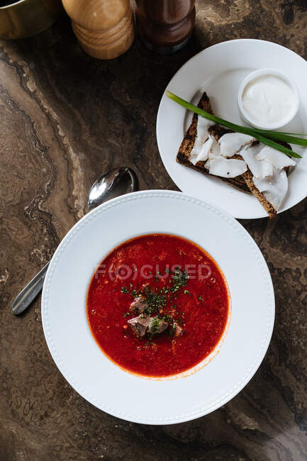 De arriba rojo sabroso apetitoso borsch espolvoreado con hierbas y pan de centeno con tocino y cebolla verde y sal en plato blanco en la mesa de madera en el restaurante - foto de stock