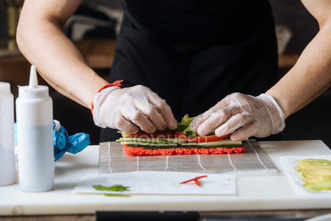 Cultivo qualificado cozinheiro em luvas descartáveis amassar recheio picante vermelho na mesa com verduras peixe e molhos na cozinha — Fotografia de Stock