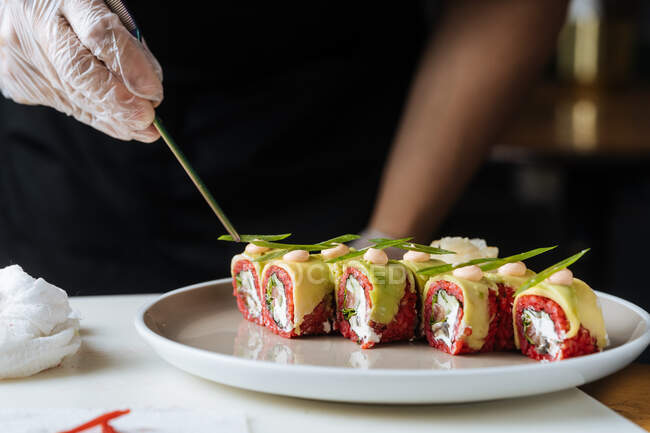 Повар в одноразовых перчатках украшает свежие вкусные суши с зеленью на белой тарелке на столе — стоковое фото