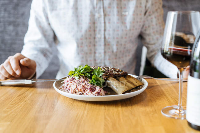 Cultivez une personne coupant des côtes de bœuf appétissantes juteuses avec une fourchette et un couteau dans une assiette avec une garniture verte à table et buvez du vin au restaurant — Photo de stock