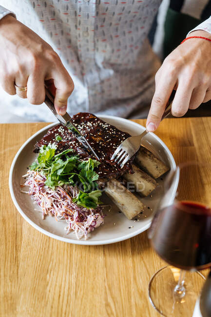 Crop person cutting juicy appetizing beef ribs with a fork and knife in plate with green garnish at table and drinking wine in restaurant — Stock Photo