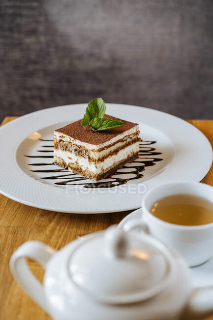 Sabroso y apetitoso tiramisú de chocolate decorado con verde y vertido con jarabe en plato blanco y té en taza blanca con tetera en la mesa en el restaurante - foto de stock