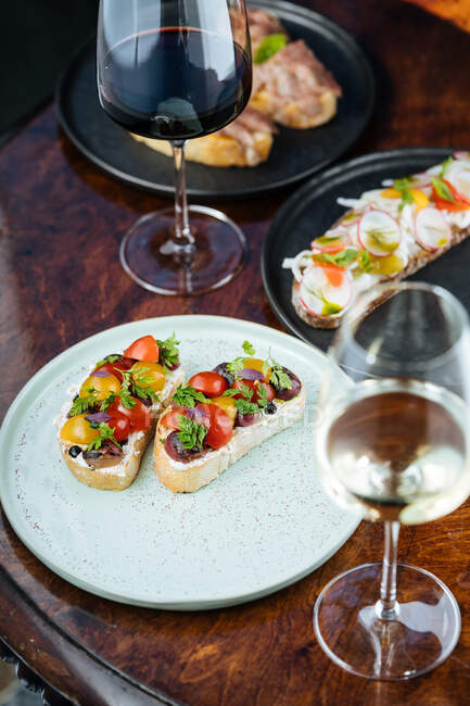 Des sandwichs colorés décorés d'en haut avec des tomates cerises jaunes rouges tranchées et des herbes sur une assiette blanche avec du vin sur la table — Photo de stock