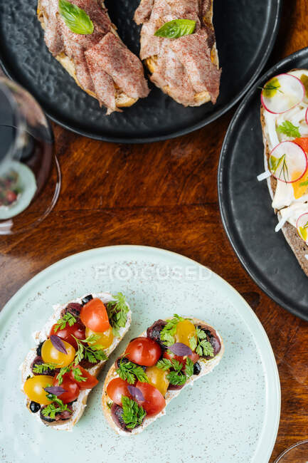 From above festive decorated colorful sandwiches with red yellow sliced cherry tomatoes and herbs on white plate with wine on table — Stock Photo