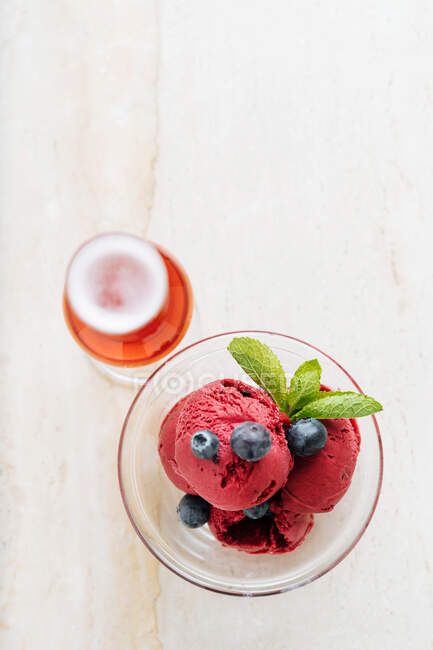 Desde arriba de cucharadas púrpuras de helado en tazón de vidrio decorado con arándanos frescos y menta y servido con vaso de bebida roja en el restaurante — Stock Photo