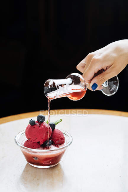 Cropped unrecognizable woman hand pouring a red beverage from a glass on purple scoops of ice cream in glass bowl decorated with fresh blueberry and mint and served in restaurant — Stock Photo