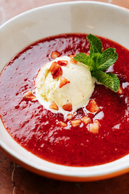 Vista superior de la cucharada de helado de vainilla en el plato con jarabe rojo decorado con pequeños trozos de fresa y menta verde fresca en el restaurante - foto de stock