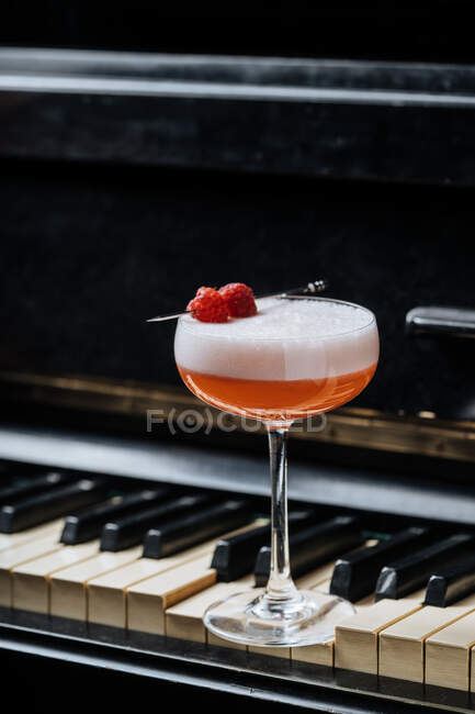 Red alcohol cocktail in stylish glass with white foam decorated with fresh raspberry on piano keys in restaurant — Stock Photo