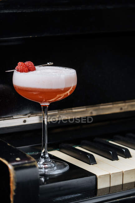 Roter Alkoholcocktail im stilvollen Glas mit weißem Schaum, dekoriert mit frischen Himbeeren auf Klaviertasten im Restaurant — Stockfoto