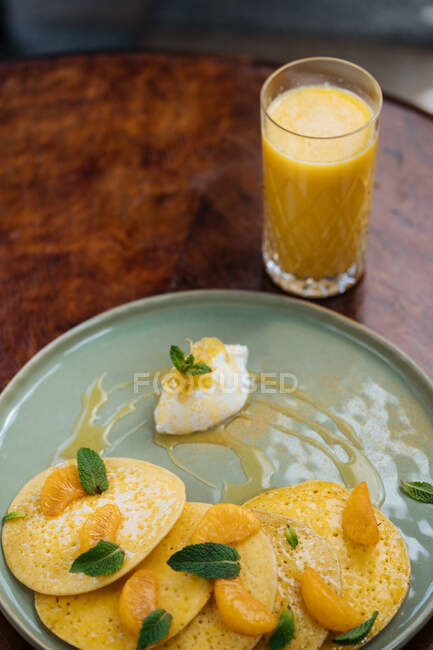 From above of plate with pancakes decorated with slices of tangerine leaves of mint served with sauce and glass of orange juice — Stock Photo