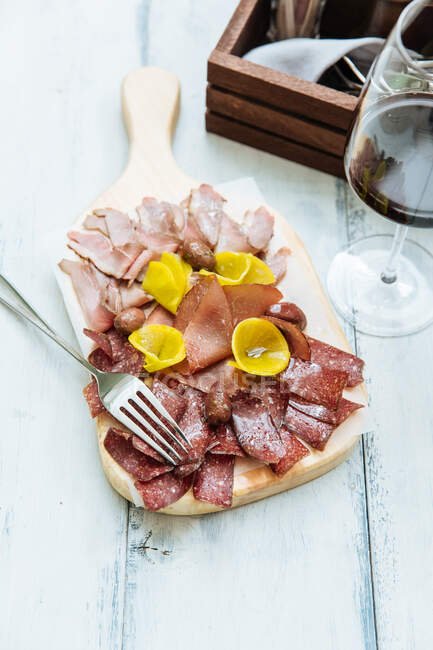 From above of slices of smoked meat different varieties creative served on wooden cutting board with metal fork and glass of red vine — Stock Photo