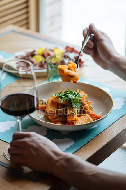 Cortada pessoa irreconhecível comendo saborosa massa vegan decorado com folhas de salada de foguete fresco e molho e beber vinho tinto em um copo em uma mesa de restaurante — Fotografia de Stock