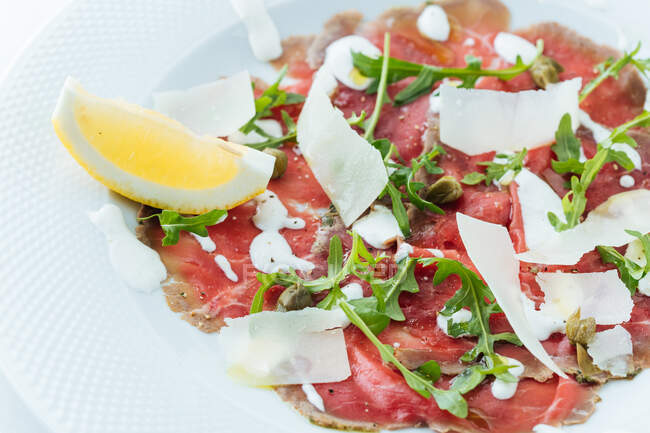 From above of slices red beef on plate with pieces of fresh lemon decorated with white sauce cheese and arugula — Stock Photo